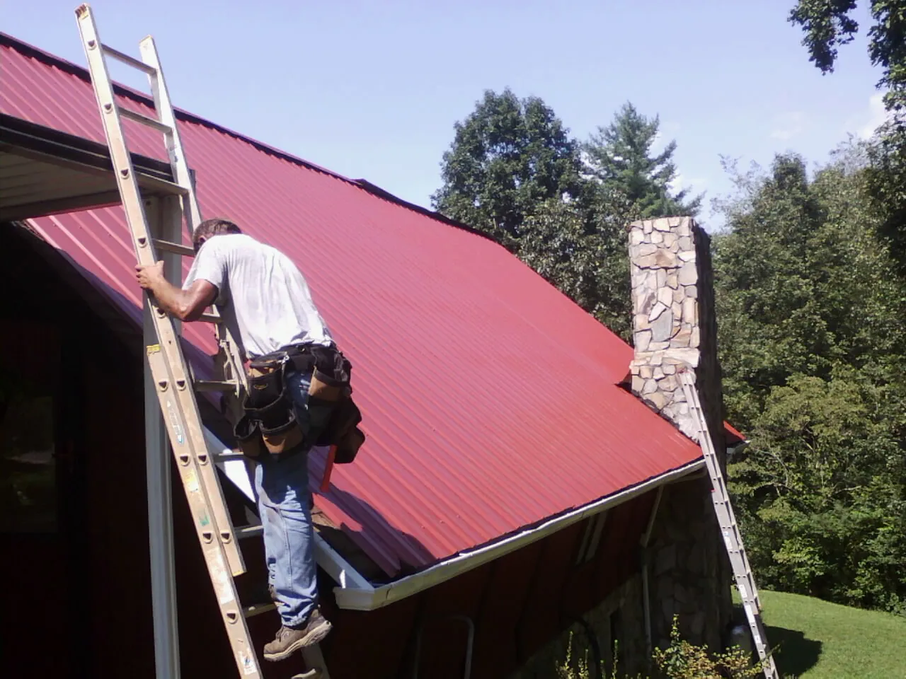 Roofer on a ladder beside a red metal roof and stone chimney — Soffit & Fascia Repair by Roofing Brothers in North Stonington
